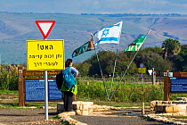 Warning sign to slow down, near woman standing by  natural methane gas upwelling, with flags, Hula Valley, Israel, November 2014.