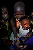 Masai family in clay hut, charging mobile phone using solar power, Mara, Kenya. March 2013.