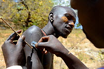 Men from the Mursi tribe decoratively scaring skin, one man scars the arm of another one by lifting the skin with an acacia spine and cutting it with a razor blade, Ethiopia, March 2015.