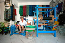Lady spinning on manually operated wheel, inside her shop, with scarves she made, Mykonos Town, Cyclades, Aegean Sea, Mediterranean, Greece, August 2007.