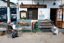 Man sitting outside a taverna, watching  whole lamb and Kokoretsi (offal sausage) cooking on a spit, in preparation for Easter Sunday meal. Spetses Island, Aegean Sea, Greece, Mediterranean, April 200...