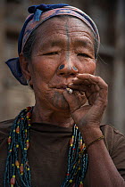 Apatani woman smoking with facial tattoos and traditional nose plugs / Yapin Hulo.These plugs are made of a cane slice. This practice was to make them look unattractive to males from other tribes. The...