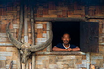 Konyak Naga Man looking out of window of house - with buffalo skull attached to the wall, Mon district.Nagaland,  North East India, October 2014.