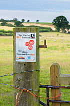 Farmers Guardian notice on field gatepost warning walkers that dogs can kill livestock, with sheep in distance. Norfolk, England UK. August.