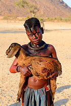 Himba girl with traditional double plait hairstyle, carrying a goat. Marienfluss Valley, Kaokoland Desert, Namibia. October 2015