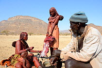 Himba man watching women cutting meat, Marienfluss Valley, Kaokoland Desert, Namibia. October 2015