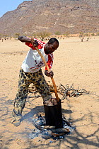 Himba man in modern western clothing cooking fresh goat meat, Marienfluss Valley, Kaokoland Desert, Namibia. October 2015