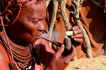 Himba woman lighting and smoking her pipe, Marienfluss Valley, Kaokoland Desert, Namibia. October 2015