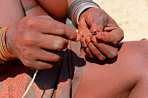 Himba woman threading metal beads to adjust her traditional 'Omohanga' foot ornament, a 21 strand layered anklet, indicating if she is married and has children, Marienfluss Valley. Kaokoland, Namibia...