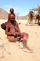 Himba woman adjusting her traditional 'Omohanga' foot ornament, a 21 strand layered anklet, indicating if she is married and has children, Marienfluss Valley. Kaokoland, Namibia October 2015