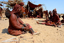 Himba woman adjusting her traditional 'Omohanga' foot ornament, a 21 strand layered anklet, indicating if she is married and has children, Marienfluss Valley. Kaokoland, Namibia October 2015