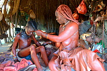 Himba woman in her hut applying Otjize (a mixture of butter,  ochre and ash) to her daughter's skin. Marienfluss Valley. Kaokoland, Namibia October 2015
