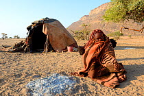 Himba woman wrapped up in a blanket next to her open fire with her mud hut in the background. Marienfluss Valley. Kaokoland, Namibia October 2015