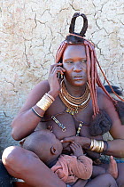 Himba woman with traditional hair and jewellery, breast feeding whilst on a mobile phone nursing her baby. Kaokoland, Namibia October 2015