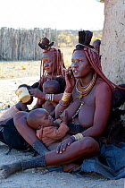 Himba woman with traditional hair and jewellery, breastfeeding whilst on a mobile phone nursing her baby. Kaokoland, Namibia October 2015