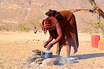 Himba woman cooking maize flour open fireplace, Marienfluss Valley, Kaokoland Desert, Namibia. October 2015