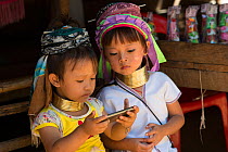 Kayan girls with neck rings looking at smartphone, Thailand. November 2015.