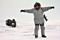 Photographer Sergey Gorshkov, standing with his arms outstretched on snow, with Musk ox (Ovibos moschatus) in the background, Wrangel Island, Far East Russia, March 2011.