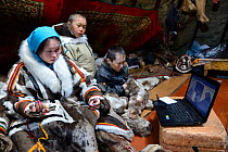 Nenet children, two boys and a teenage girl, watching laptop inside tent. The girl is sewing reindeer skin. Yar-Sale district. Yamal, Northwest Siberia, Russia. April  2016.