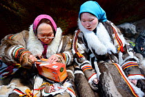 Two generations of Nenet women, an older woman and teenage girl, sewing and wearing traditional coats made of reindeer skin. Yar-Sale district, Yamal, Northwest Siberia, Russia. April 2016.