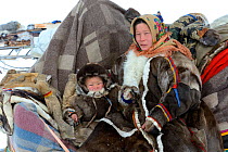 Nenet herder on sled with young boy during spring migration wearing traditional coat made with reindeer skin. Yar-Sale district. Yamal, Northwest Siberia, Russia. April 2016.