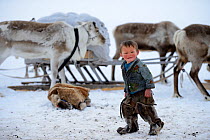 Young Nenet boy at camp wearing traditional winter boots made with reindeer skin. Reindeer (Rangifer tarandus) and sled in background. Yar-Sale district. Yamal, Northwest Siberia, Russia. April  2016.