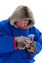 Nenet reindeer herder in traditional malitsa (reindeer skin coat) with smartphone. Yar-Sale district, Yamal, Northwest Siberia, Russia. April  2016.