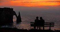 Elderly tourists sitting on bench looking at the Porte D'Aval, a natural arch in the chalk cliffs at Etretat at sunset, Cote d'Albatre, Upper Normandy, France, October 2015.