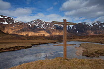 Signpost for the Affric Kintail Way long distance footpath beside the River Affric,  moorland with snow covered mountains. Glen Affric National Nature Reserve, Highlands, Scotland, UK, April 2015.
