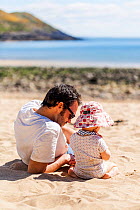 RF - Father and daughter, aged 12 months, playing with seaweed on Langland Beach, Gower, Wales. Model released. April. (This image may be licensed either as rights managed or royalty free.)