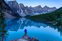 Person standing on rocks by looking over lake in the Rocky Mountains, with surrounding forest and kayaks stored, Alberta, Canada. July.