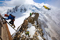 Climbers on the Cosmiques Arete, climbing the ladder to access the cable car station, and a base jumper in a wing suite flying past. Mont Blanc from the Aiguille Du Midi above Chamonix, France, Septem...