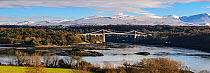 Menai Suspension Bridge, designed by Thomas Telford, viewed from Anglesey across Menai Strait, with snow capped hills in background. North Wales, UK. December 2017