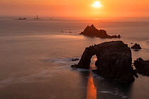 Sunset through Enys Dodnan Arch, with the Armed Knight rock and Longships lighthouse in the distance, Land's End, Cornwall, UK. May 2018.