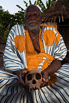 Local manon man in traditional robe with Chimpanzee (Pan troglodytes verus) skull,  Bossou, Republic of Guinea. December 2012.