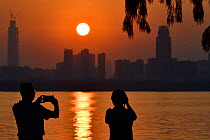 Two people taking a picture of the sunset skyline of Wuhan, East Lake Greenway park, Wuhan, Hubei, China. June 2018