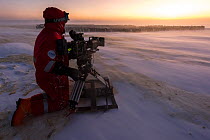 Cameraman Lindsay McCrae filming huddling Emperor penguin (Aptenodytes forsteri) colony during winter storm and polar night, for BBC Dynasties Penguin programme. Atka Bay, Antarctica. June 2017.