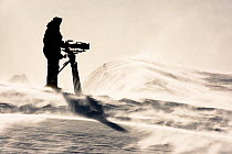 Cameraman Lindsay McCrae on sea ice filming frozen landscape and icebergfor BBC Dynasties Penguin programme. Atka Bay, Antarctica. October 2017.