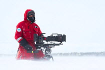 Camerman Lindsay McCrae with film camera in front of Emperor penguin (Aptenodytes forsteri) breeding colony,during winter storm, on location for BBC Dynasties Penguin programme. Atka Bay, Antarctica....