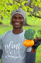 Vegan man holding broccoli and a yellow pepper, wearing a &#39;plant powered&#39; t-shirt. North London, England, UK. March 2019.