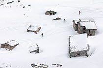 Group of snowshoe hikers passing snow covered mountain refuges in spring. These refuges are used in summer by herders to sleep while watching for their cattle, Gran Paradiso National Park, Aosta Valle...