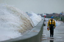 Waves crashing against the sea wall with a coast guard walking along it, Rhyl, Wales. January.