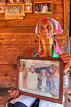 Elderly Kayan Lahwi woman (80 years old) with brass neck coils and traditional clothing sitting on her home deck and showing pictures of her when she was young. The black and white picture shows her a...