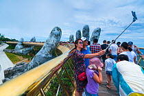 Tourists taking selfie on Golden Bridge with giant fiberglass hand appearing to hold up bridge, Sun World Ba Na Hills, Danang, Vietnam.