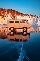 Minibus parked on ice with rock cliffs on shore, reflected in Lake Baikal, Siberia, Russia. February 2019.