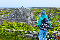 Cyclist looking at view of Seven Churches and countryside beyond. Inishmore, Aran Islands, County Galway, Republic of Ireland. May 2011.