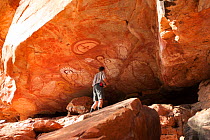Tourist looking at Wandjina style Aboriginal rock art, depiction of people and fish. Raft Point, Doubtful Bay, The Kimberley, Western Australia. 2016.