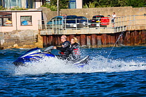 Man and woman on jet ski with harbour wall beyond. Poole Harbour, Dorset, England, UK. September 2018.