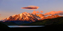 Light at sunrise on towers and Central Massif of Torres del Paine National Park, lenticular cloud above mountains, Laguna Amarga in foreground. Torres del Paine National Park, Patagonia, Chile. November 2018.