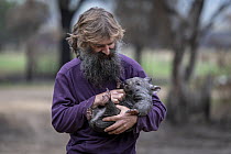 Portrait of Joseph Henderson holding a male bare-nosed wombat (Vombatus ursinus(. He and his partner, Rena, had to evacuate the wildlife (wombats, kangaroos and possums) from their home and wildlife s...